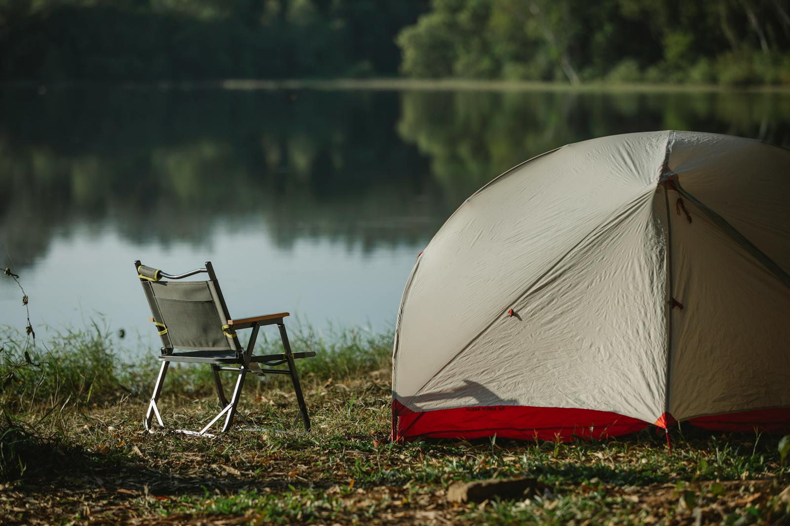 Tent and folding chair on grass coast against trees reflecting in lake in soft sunlight